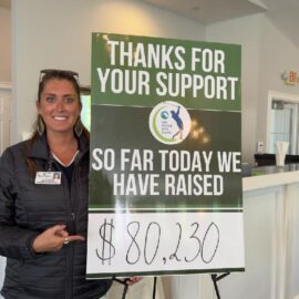 LeeAnn Clark Moore stands next to a sign that reads, "Thanks for your support. So far today we have raised $80,230." She is smiling and pointing at the amount raised on the sign.