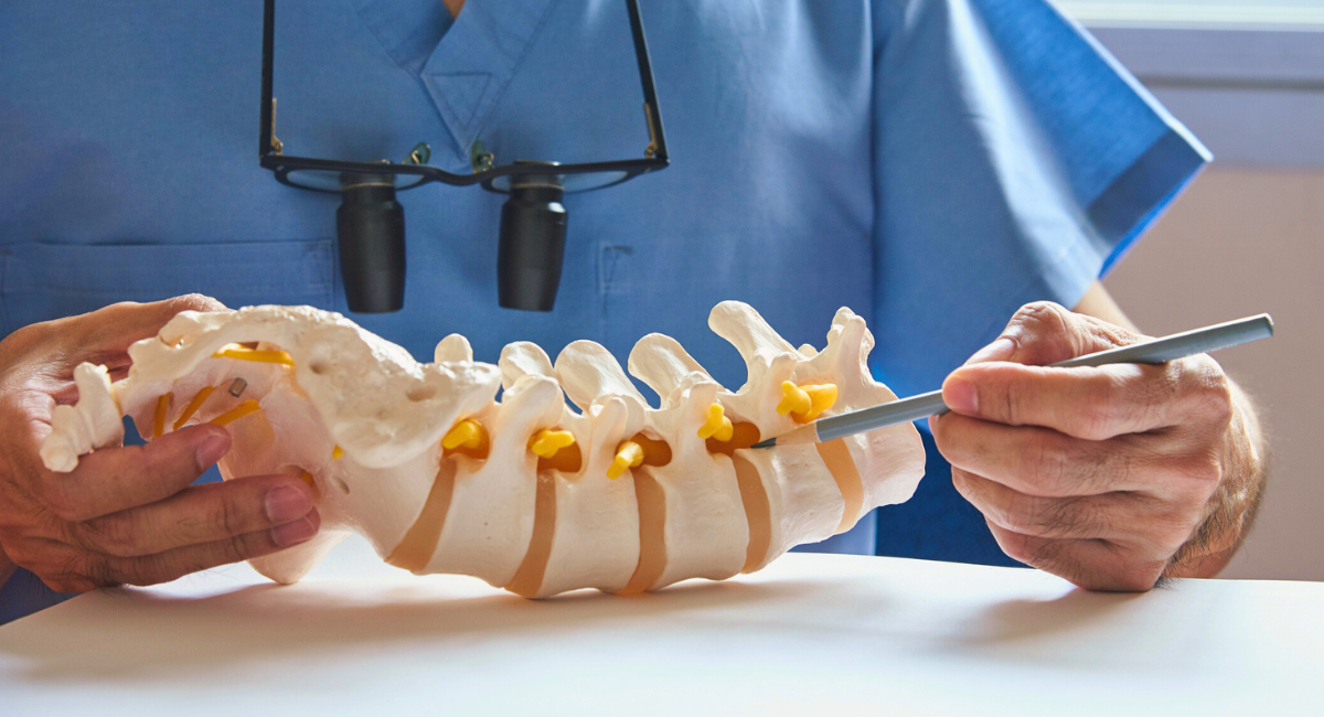 A healthcare professional wearing magnifying surgical glasses points to a model of the human spine, illustrating key details about spinal anatomy and care.