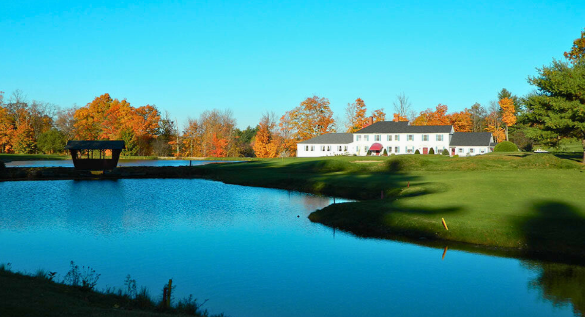 Scenic golf course view with a pond and a small covered bridge in the foreground, autumn trees with orange foliage in the background, and a large white clubhouse with red awnings under a clear blue sky.