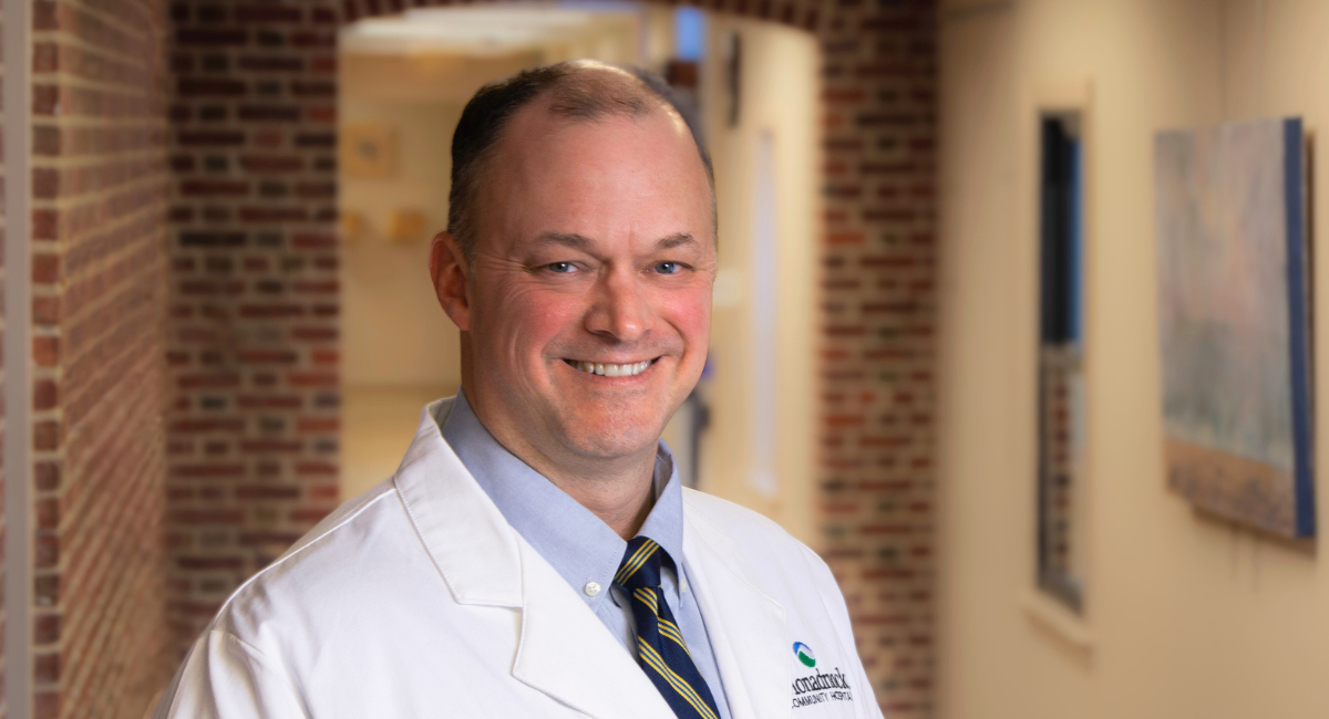 Dr. Michael Gilbert, wearing a white medical coat with the Monadnock Community Hospital logo, smiles in a warmly lit hallway with brick walls and framed artwork.