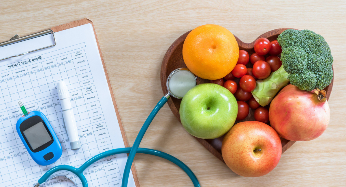 A heart-shaped wooden bowl filled with fresh fruits and vegetables, including an apple, orange, broccoli, and tomatoes, next to a stethoscope, blood glucose monitor, and a blood sugar tracker clipboard. The image represents a healthy lifestyle and management tools for people living with diabetes, emphasizing the importance of nutrition and regular monitoring, aligning with a diabetes support group's focus on wellness