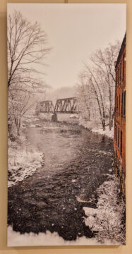 Photo on canvas snowy scene of river with trees brick building on right metal bridge ahead