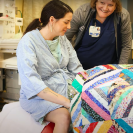 A pregnant woman sitting on a hospital bed, covered with a colorful quilt, smiling, and being assisted by a nurse in the birthing suite at Monadnock Community Hospital