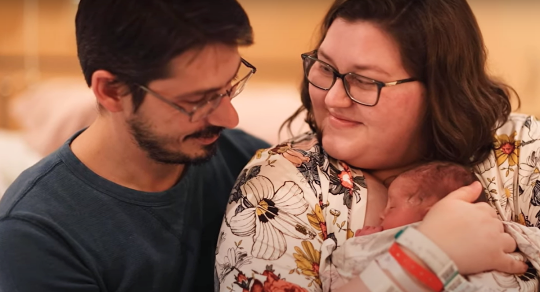 A happy couple holding their newborn baby. The mother is smiling warmly while holding the baby close, and the father is looking at both lovingly