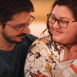 A happy couple holding their newborn baby. The mother is smiling warmly while holding the baby close, and the father is looking at both lovingly