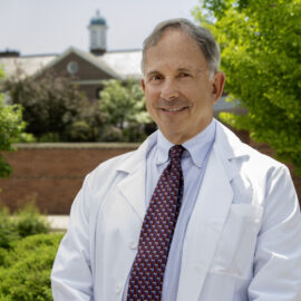 Dr. Jonathan Krant stands outside, wearing a white lab coat and a patterned tie, with a professional demeanor. The background features a well-maintained garden with lush greenery and a brick building topped with a cupola. Dr. Krant is smiling, conveying a sense of warmth and approachability, representing his role as a dedicated rheumatologist at Monadnock Rheumatology Associates.