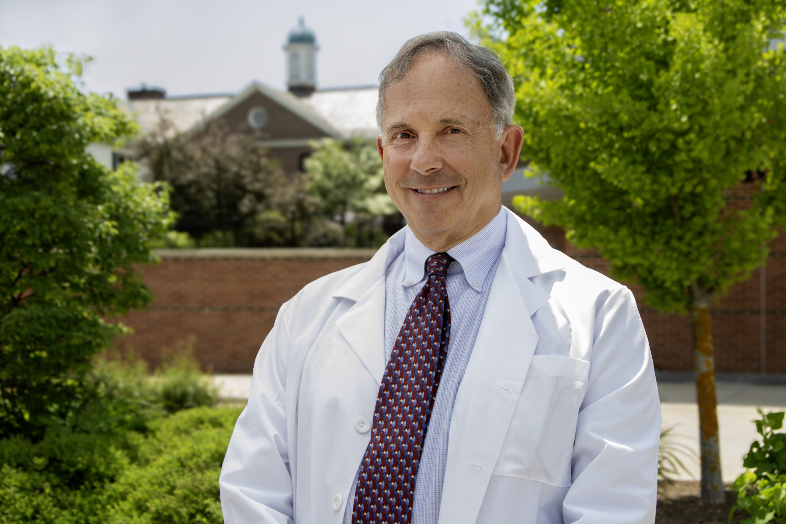 Dr. Jonathan Krant stands outside, wearing a white lab coat and a patterned tie, with a professional demeanor. The background features a well-maintained garden with lush greenery and a brick building topped with a cupola. Dr. Krant is smiling, conveying a sense of warmth and approachability, representing his role as a dedicated rheumatologist at Monadnock Rheumatology Associates.