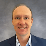 Headshot of a man with short light hair, wearing a dark blazer and light blue striped shirt, smiling in front of a plain background