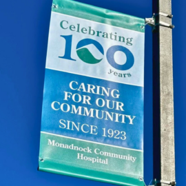 A colorful banner against a bright blue sky, celebrating Monadnock Community Hospital's 100th anniversary with the slogan "Caring for Our Community Since 1923."