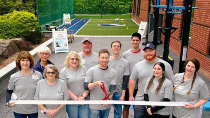 A group of people standing in front of an outdoor rehab space, smiling and holding a ribbon for a ribbon-cutting ceremony. The rehab space includes various equipment and a blue court in the background.