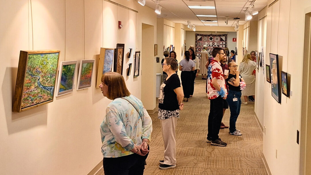 A hallway in Monadnock Community Hospital displaying various pieces of artwork. Several people are observing the art, including paintings and framed pictures, which are hung on the walls. The atmosphere is lively, with individuals engaged in conversation and appreciating the exhibits. The lighting is warm, enhancing the colors and details of the artwork. The setting is inviting, showcasing the hospitals support for local artists and the communitys cultural engagement.