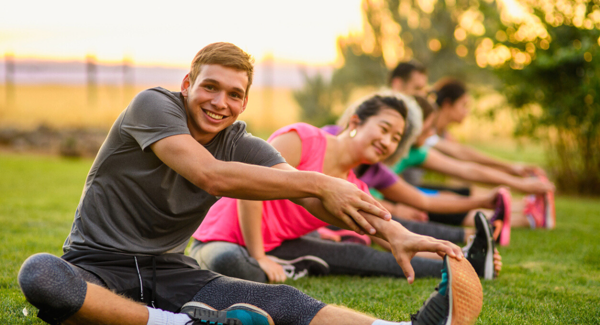 People stretching outdoors in a fitness class on a grassy field, smiling and enjoying the activity together at sunset