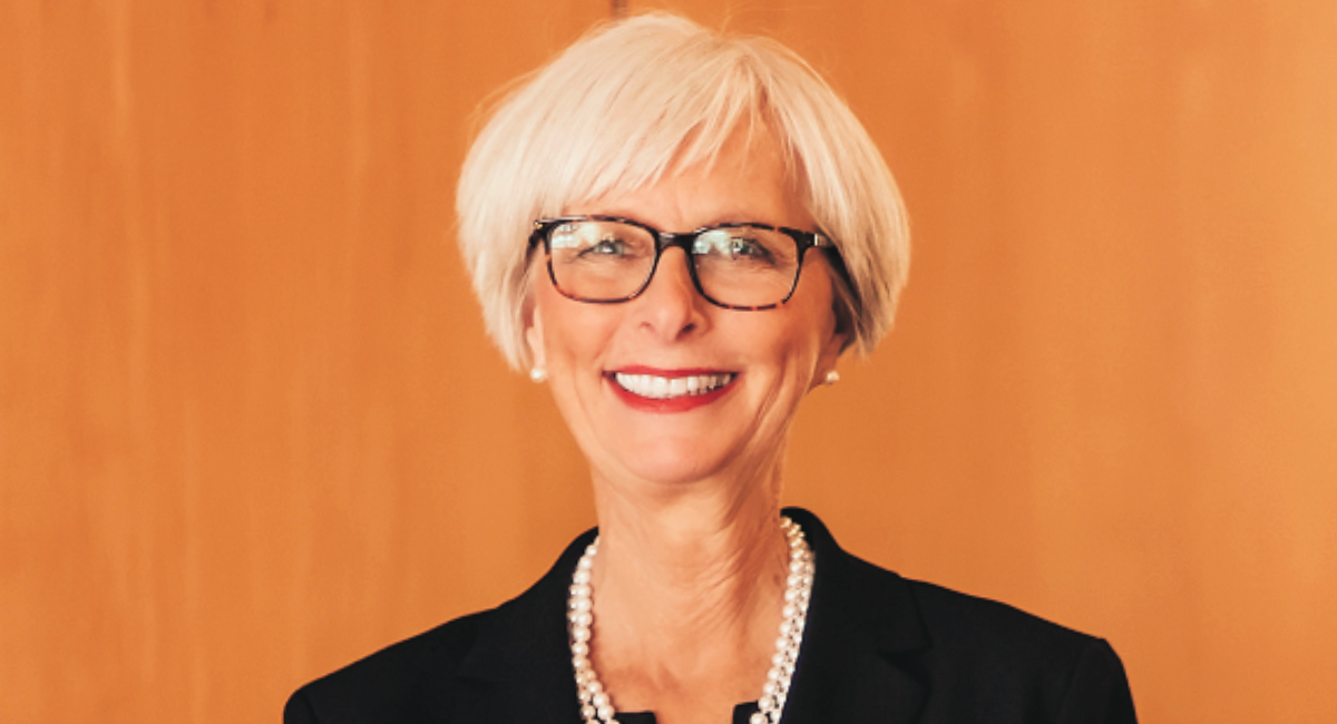 Smiling woman with short white hair and glasses, dressed in professional attire, standing in front of a wooden background