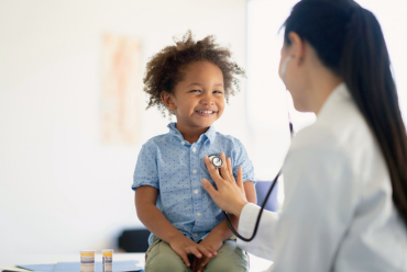 A smiling young child in a blue shirt having their heartbeat checked by a healthcare provider with a stethoscope in a bright, clean medical office.