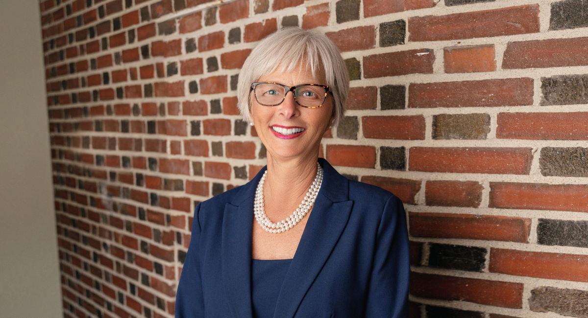 Cynthia K. McGuire, President & CEO of Monadnock Community Hospital, standing against a brick wall, smiling and wearing a navy blue suit with a pearl necklace.