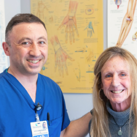 Dr. Vache Hambardzumyan, wearing blue scrubs, smiles alongside a female patient in an examination room, with anatomical charts in the background.