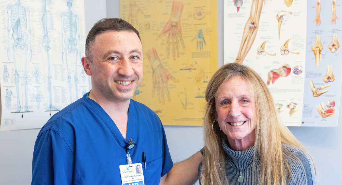 Dr. Vache Hambardzumyan, wearing blue scrubs, smiles alongside a female patient in an examination room, with anatomical charts in the background.