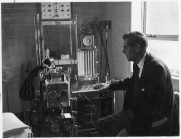 A black-and-white photograph of Dr. Chandler sitting beside a piece of medical equipment. He is dressed in a suit and appears focused on the device, reflecting the medical technology and professional attire of the period.