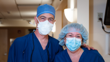 Two medical professionals in blue scrubs and surgical masks, smiling at the camera inside Monadnock Community Hospital.