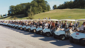 A row of golf carts lined up with participants ready to start a tournament, set against a backdrop of green hills and trees.