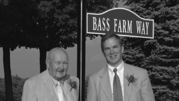 A black-and-white photo of Herb Nilson and Peter Gosline standing next to a sign that reads "Bass Farm Way," both dressed in light-colored suits and smiling.