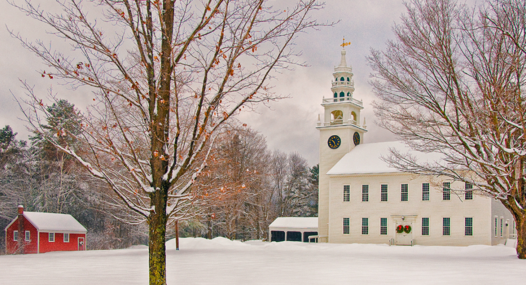 The historic Jaffrey Meetinghouse in Jaffrey, covered in snow with a backdrop of winter trees, showcasing classic New England architecture