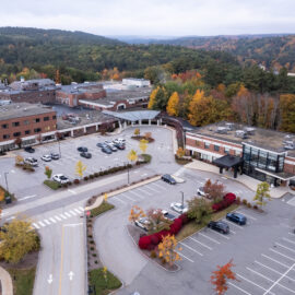 An aerial view of Monadnock Community Hospital surrounded by a lush forest with autumn foliage. The hospital complex consists of multiple interconnected brick buildings with parking lots and landscaped areas. Cars are parked in the lots, and the main entrance is marked by a curved driveway and canopy. The backdrop features rolling hills covered in vibrant fall colors, creating a picturesque and serene setting.