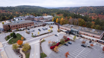 An aerial view of Monadnock Community Hospital surrounded by a lush forest with autumn foliage. The hospital complex consists of multiple interconnected brick buildings with parking lots and landscaped areas. Cars are parked in the lots, and the main entrance is marked by a curved driveway and canopy. The backdrop features rolling hills covered in vibrant fall colors, creating a picturesque and serene setting.