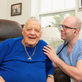 A clinician, dressed in blue scrubs, attentively listens to an elderly male patient sitting in a recliner at home, showcasing the Mobile Integrated Health program.