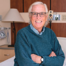 An elderly male patient, smiling, sits on a hospital bed in a well-lit room, with his arms crossed.