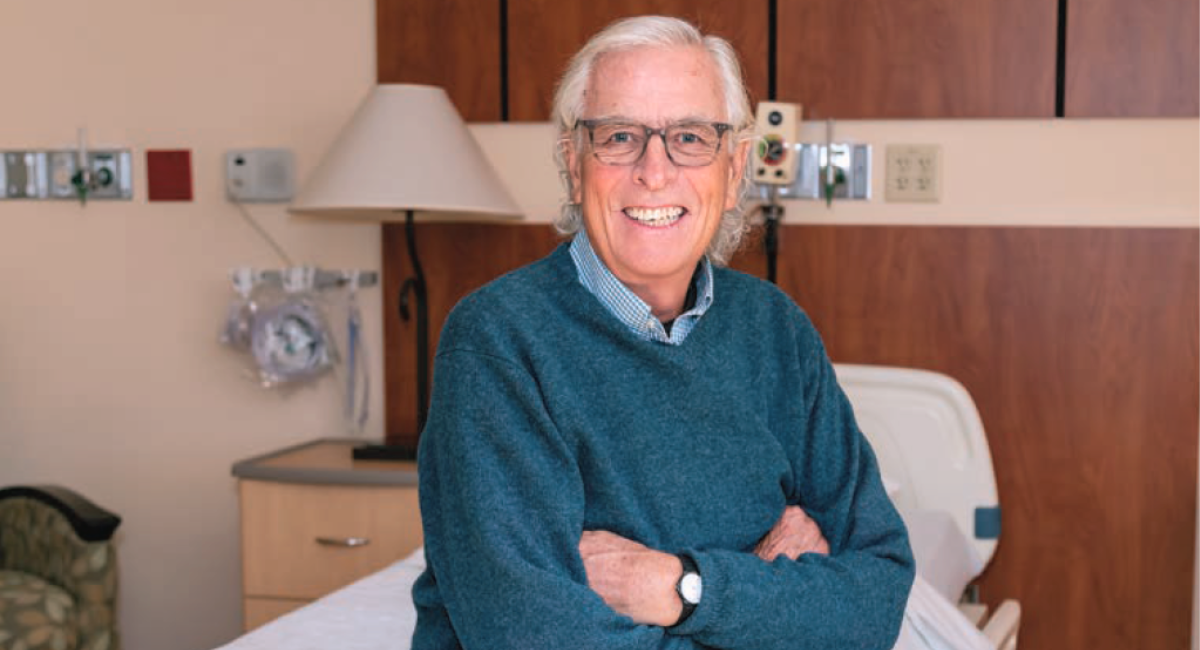 An elderly male patient, smiling, sits on a hospital bed in a well-lit room, with his arms crossed.