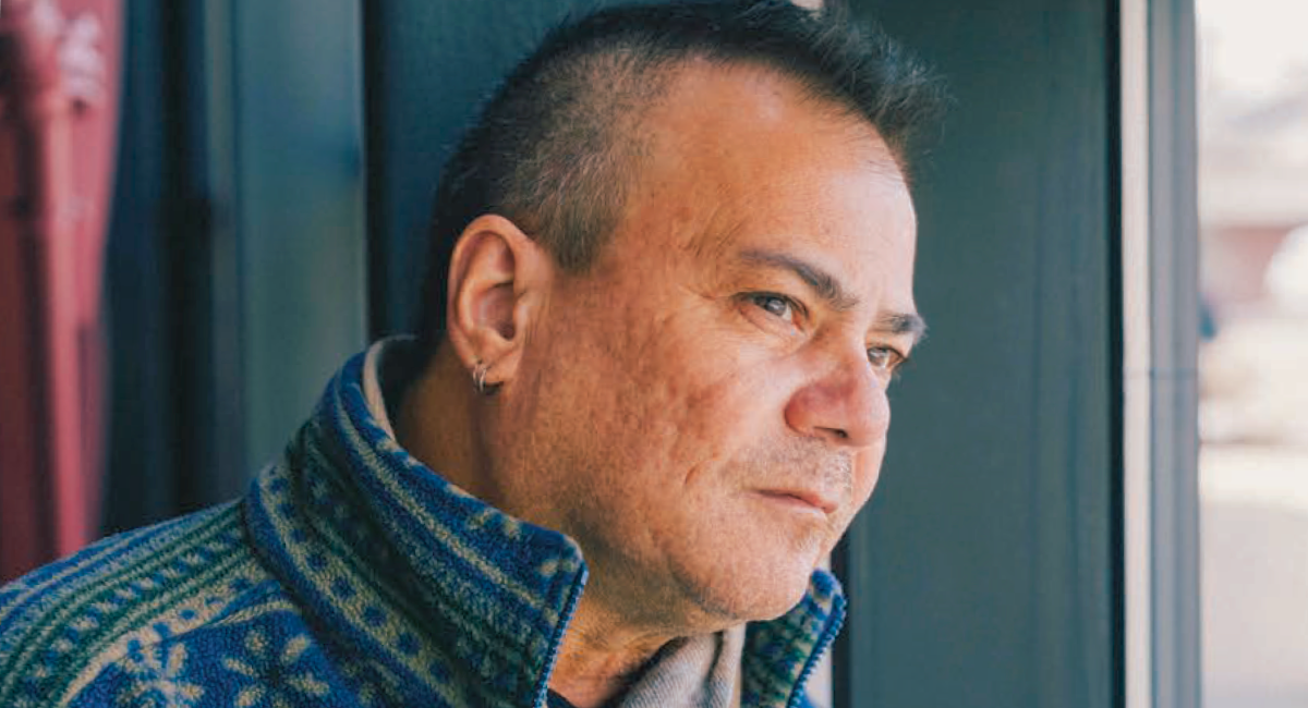 A thoughtful male patient gazing out of a window, wearing a blue and green patterned sweater.