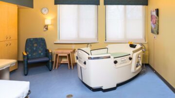 A color photograph of a birthing room featuring a birthing tub, comfortable chair, and medical equipment. The room is designed to provide a comfortable and soothing environment for childbirth, with soft lighting and modern amenities.