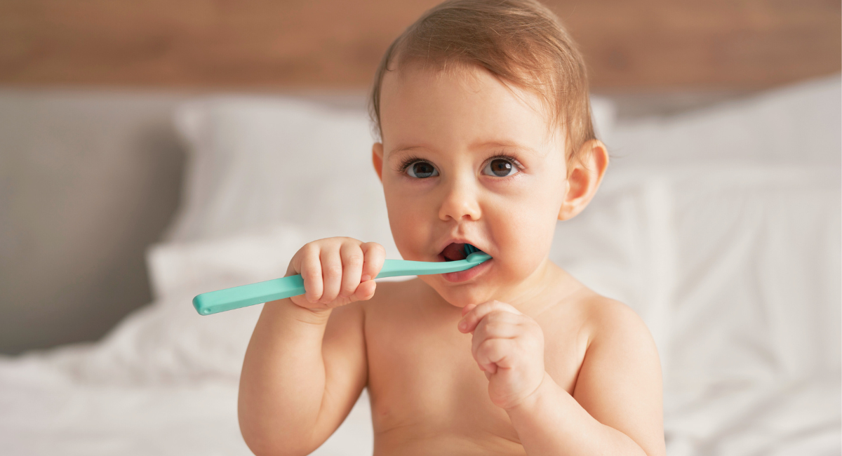 A baby is sitting on a bed, holding and chewing on a turquoise toothbrush, with a white towel partially wrapped around them