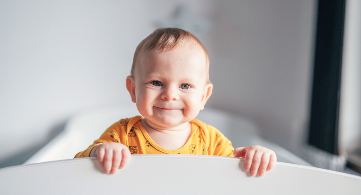 A smiling baby in a yellow onesie with animal prints is standing in a white crib, looking directly at the camera.