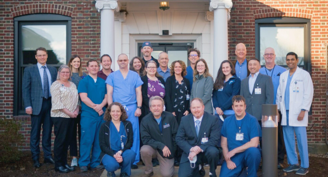 A diverse group of healthcare professionals, including doctors in white coats and staff in blue scrubs, standing in front of a brick building with white columns. They are smiling and appear welcoming, representing the comprehensive health services team at Monadnock Community Hospital.