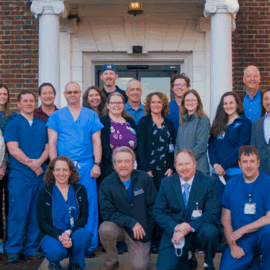 A diverse group of healthcare professionals, including doctors in white coats and staff in blue scrubs, standing in front of a brick building with white columns. They are smiling and appear welcoming, representing the comprehensive health services team at Monadnock Community Hospital.