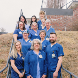 A group of twelve healthcare professionals in blue scrubs and work attire, standing on an outdoor staircase. They are smiling and looking at the camera, showcasing the collaborative and friendly team at Monadnock Community Hospital