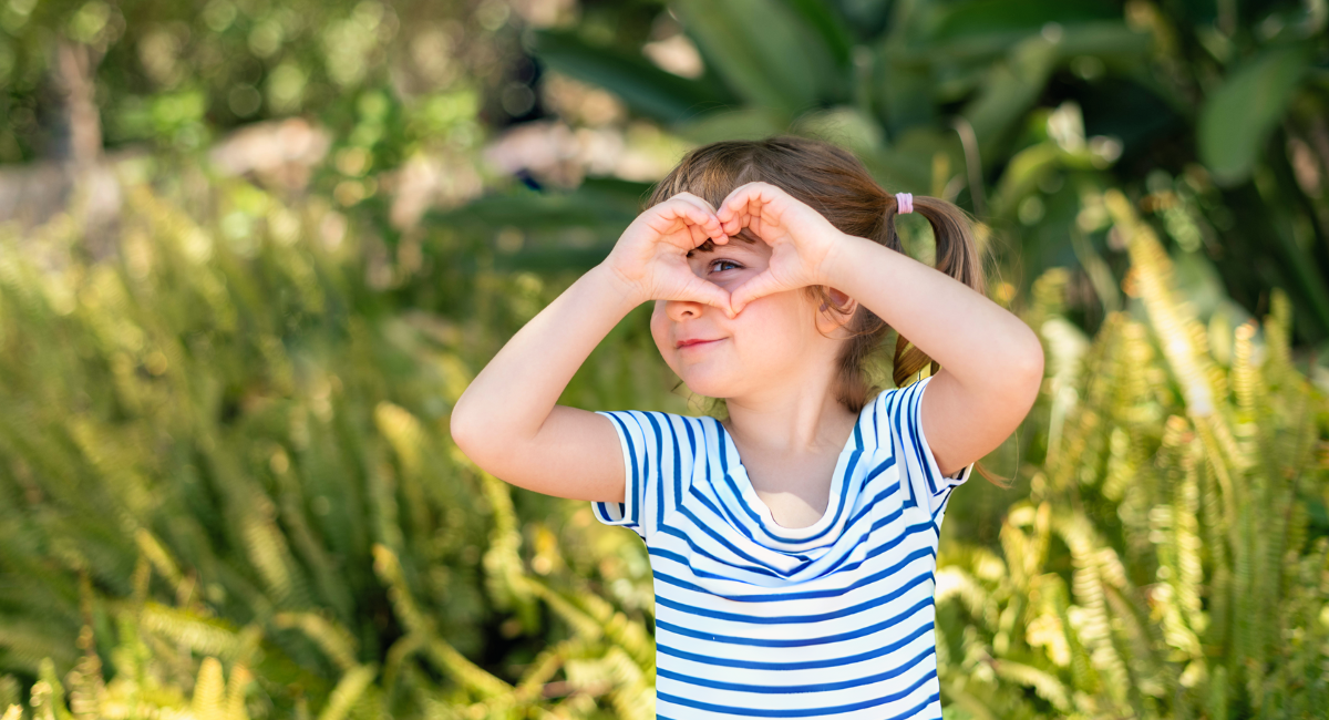 A young child in a black and white striped shirt is standing outdoors, making a heart shape with their hands around their eyes, smiling