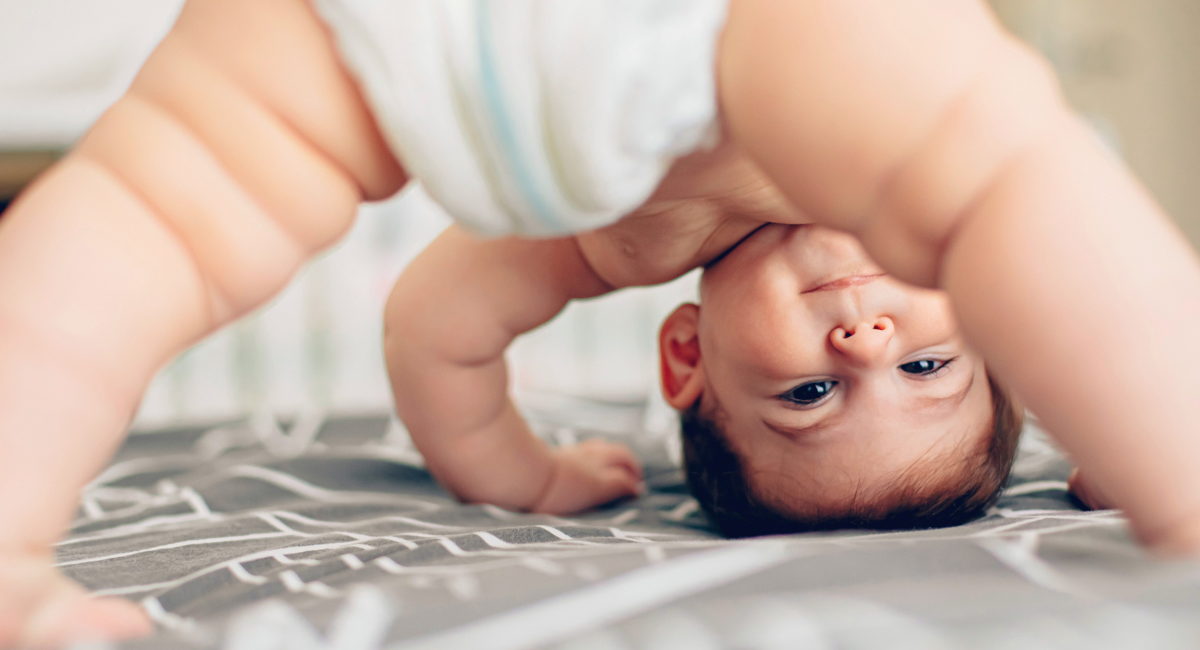A baby in a diaper is upside down on a bed, looking through their legs and smiling