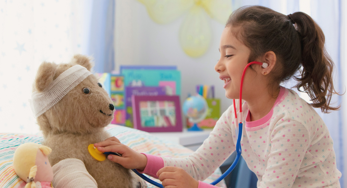 A young child, wearing polka dot pajamas, is smiling as they play doctor with a teddy bear, using a stethoscope to check the bear's heartbeat. The teddy bear has bandages on its head and arm