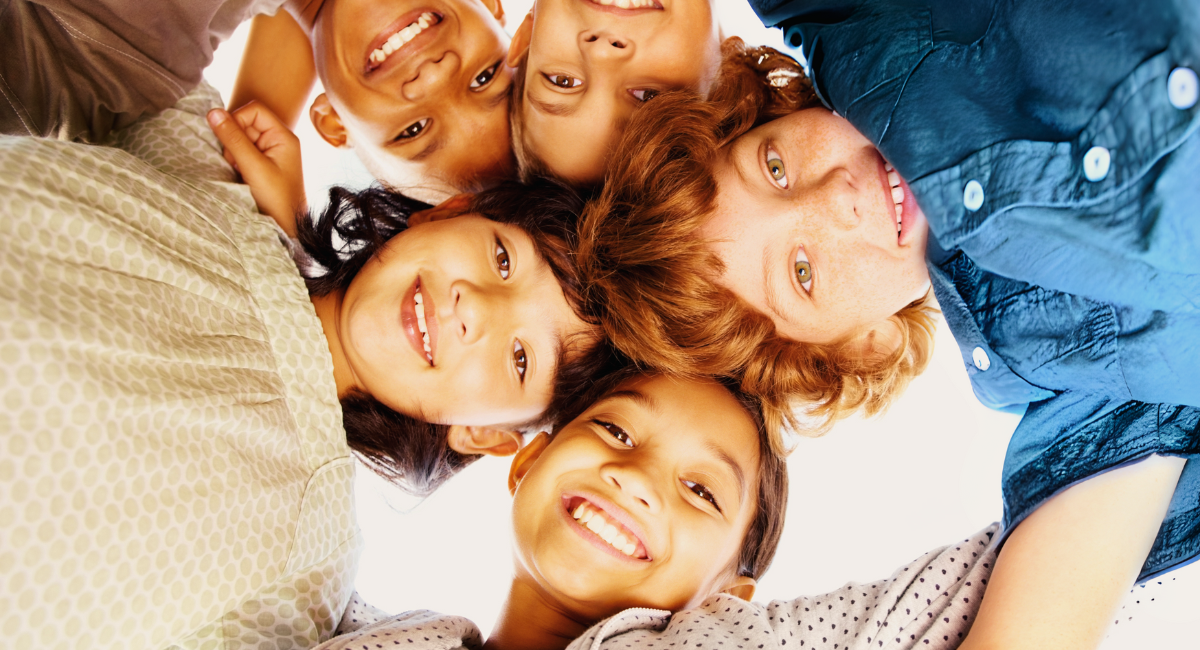 Five children are huddled together, smiling and looking down at the camera. They are wearing casual clothing and appear happy and close-knit