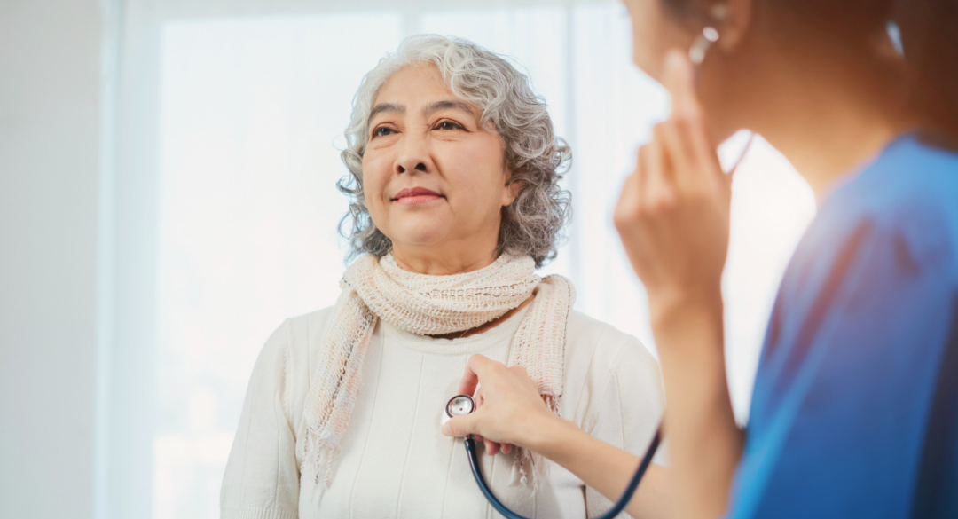 An older woman with curly gray hair wearing a light scarf and sweater, receiving a check-up from a healthcare professional. The professional is holding a stethoscope to the woman's chest in a bright, softly lit room, conveying a sense of care and attentiveness.