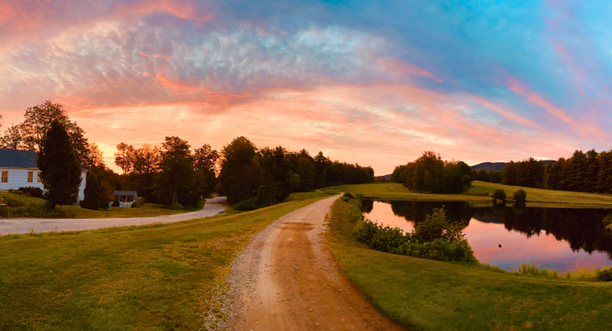 A serene dirt golf cart path curves alongside a peaceful pond under a vibrant sunset sky, surrounded by lush greenery and trees. A white house is visible on the left, nestled among the trees, adding a charming touch to the tranquil rural landscape.