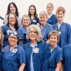 Group of Monadnock Community Hospital healthcare professionals standing together outside in front of a brick building, all wearing blue uniforms and smiling at the camera.