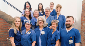 Group of Monadnock Community Hospital healthcare professionals standing together outside in front of a brick building, all wearing blue uniforms and smiling at the camera.