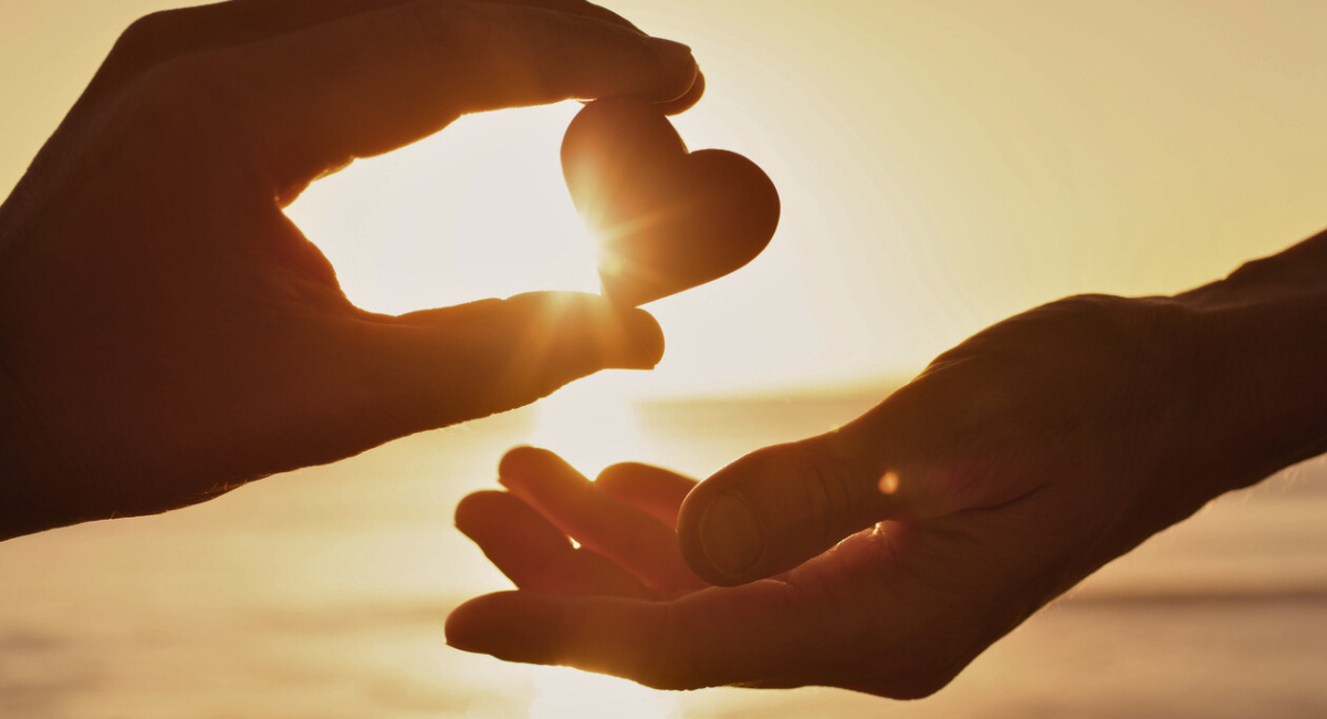 Two hands reaching towards each other at sunset, one holding a heart-shaped object illuminated by the sun's rays, symbolizing compassion and community support