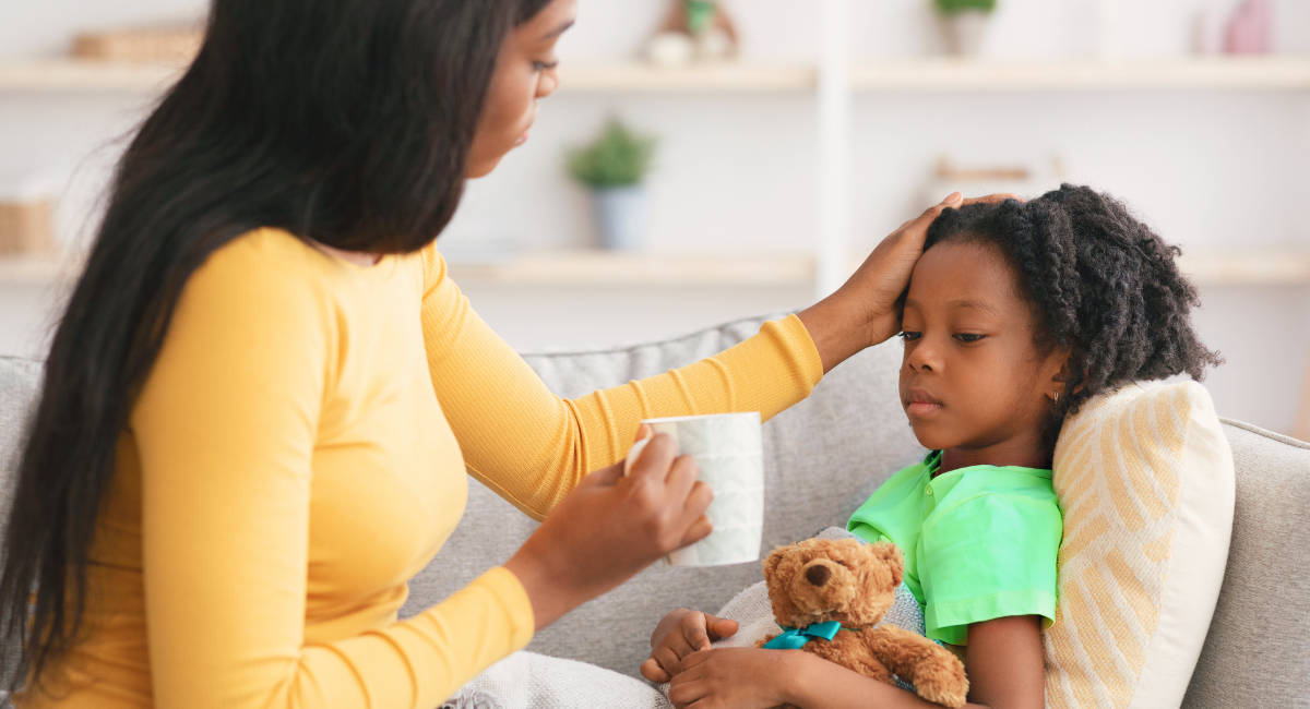 A parent wearing a yellow shirt, is sitting on a couch and checking the temperature of their child who is holding a teddy bear and wrapped in a blanket.