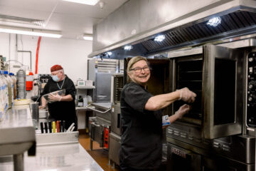 A color photograph of a modern hospital kitchen with two staff members working. One is smiling at the camera while opening an oven, and the other is preparing food in the background. The kitchen is equipped with stainless steel appliances and cooking utensils, reflecting a contemporary and well-equipped culinary environment.