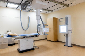 A color photograph of a modern X-ray room with advanced imaging equipment, including a large X-ray machine and a patient table. The room is clean and well-organized, reflecting current medical imaging technology.